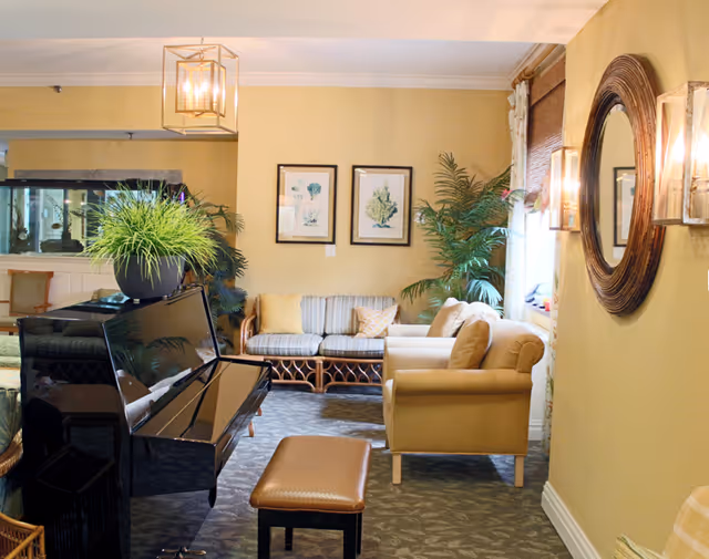 A cozy living room area with a black upright piano and matching bench in the foreground. Behind the piano, there is a wicker sofa with striped cushions and several throw pillows. To the right, there is a beige armchair. The walls are painted a warm beige color and decorated with two framed botanical prints and a large round mirror with wall sconces on either side. There are also several green plants adding a touch of nature to the space.