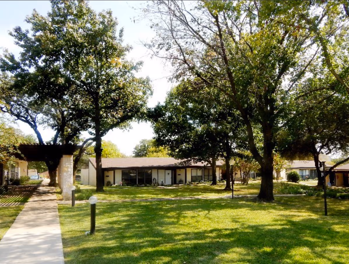A sunny outdoor area at Lake Forest Village featuring a well-maintained grassy lawn, several mature trees providing shade, a paved walkway, and low-rise residential buildings in the background.
