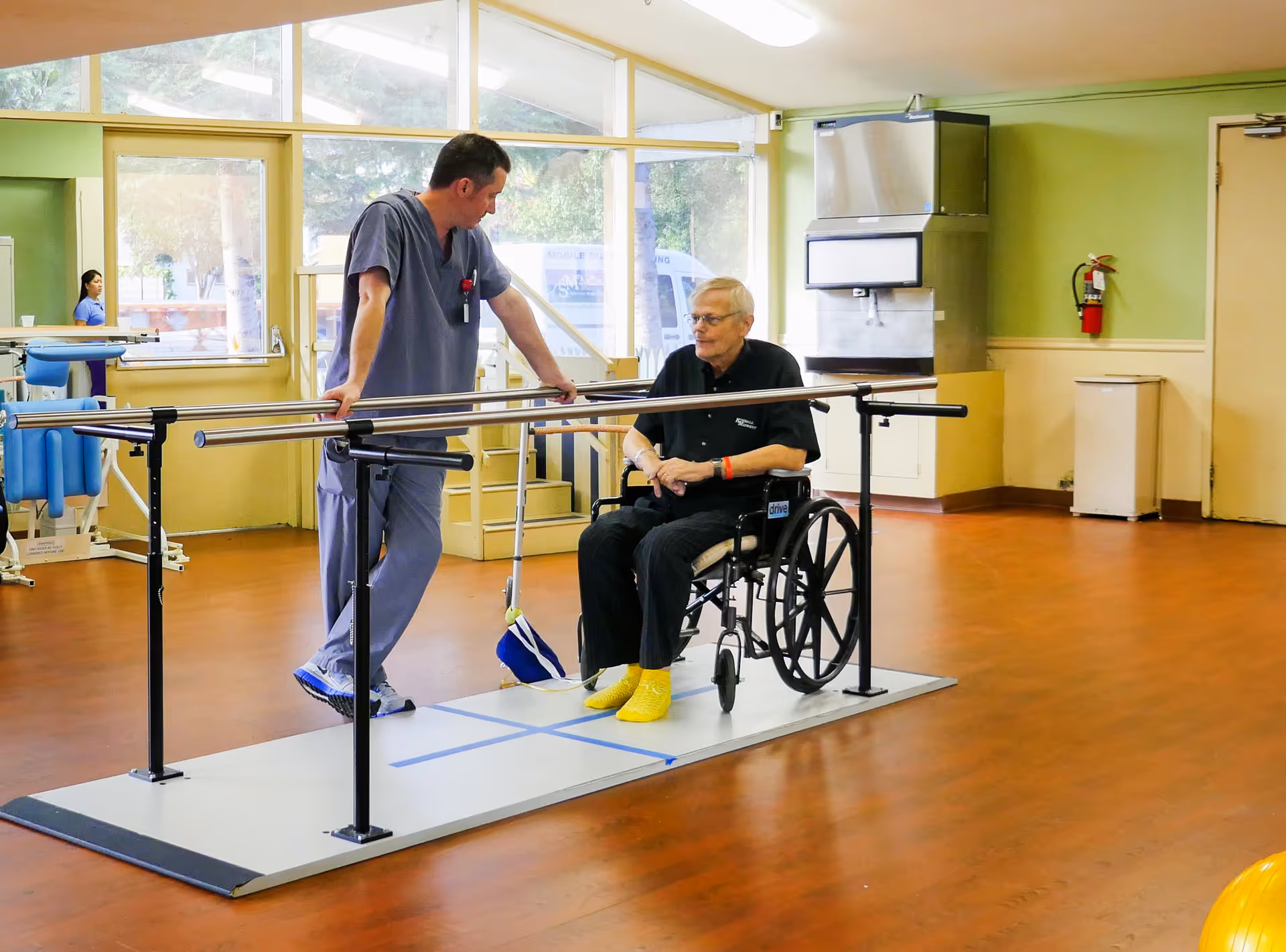 A healthcare professional in scrubs is assisting an elderly man in a wheelchair in a rehabilitation center room with wooden floors and large windows. The man is positioned on a platform with parallel bars, likely used for physical therapy exercises. The room has light green walls, a fire extinguisher, and medical equipment in the background.