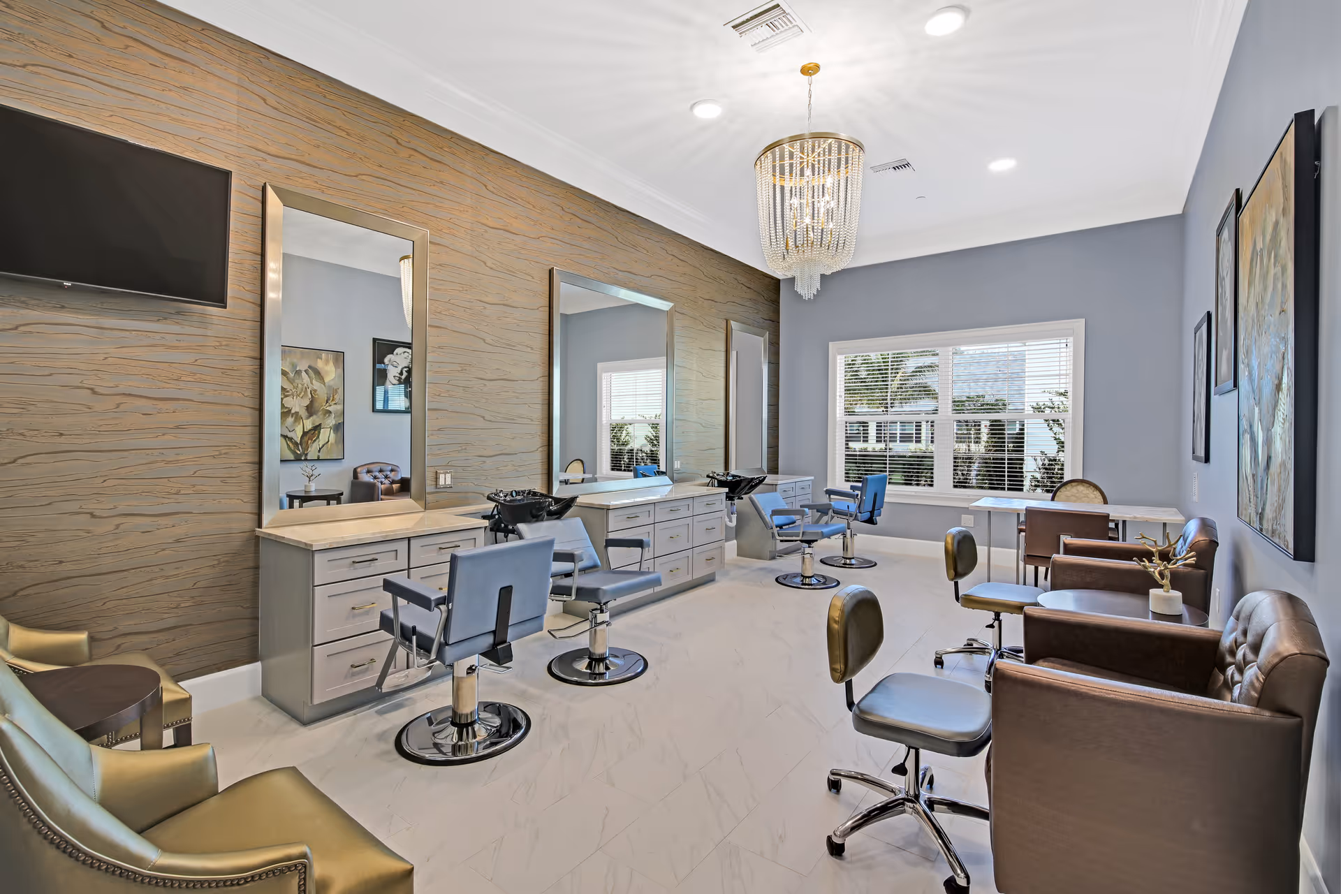 A bright and modern salon room in a senior living facility featuring three styling chairs in front of large mirrors with built-in cabinets, a flat-screen TV mounted on the wall, comfortable seating including leather armchairs and stools, a chandelier hanging from the ceiling, and large windows letting in natural light.