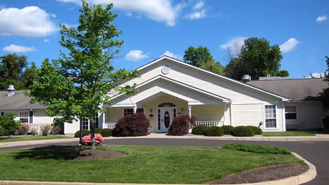 Single-story building with light yellow siding and a gray roof, featuring a covered entrance with white columns. The building is surrounded by a well-maintained lawn, bushes, and a tree in the front yard under a blue sky with some clouds.