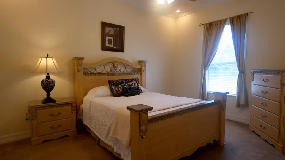 A cozy bedroom with a wooden bed frame, white bedding, and two decorative pillows. There is a nightstand with an ornate lamp on the left side of the bed, a window with beige curtains on the right wall, and a wooden dresser with multiple drawers next to the window. A framed picture hangs on the wall above the bed.