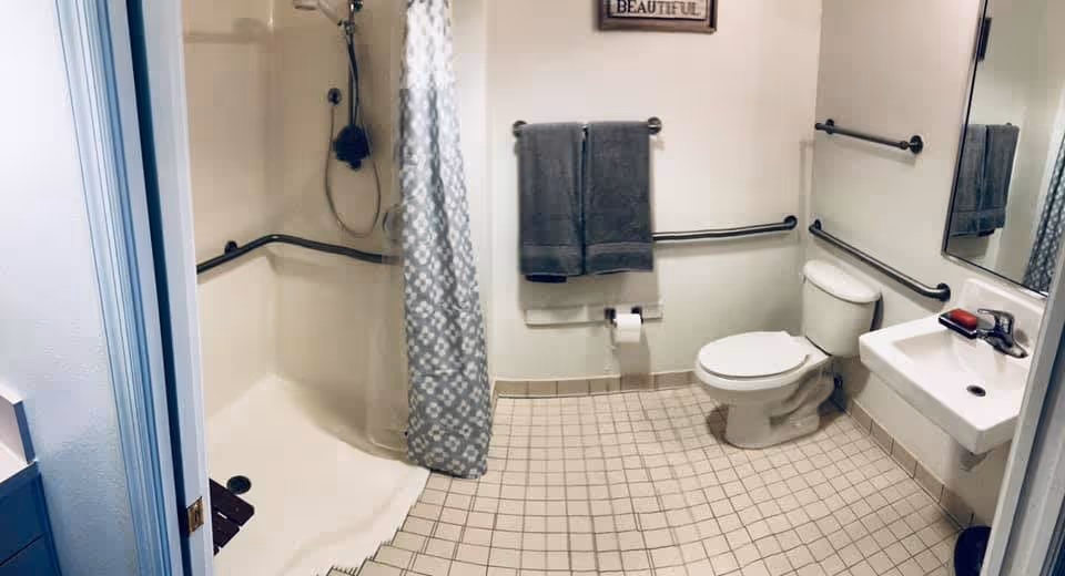 A clean and accessible bathroom with beige tiled floor, a white toilet, a white sink with a mirror above it, and a walk-in shower with a gray patterned curtain. There are multiple grab bars on the walls for support, two gray towels hanging on a towel rack, and a small sign on the wall above the towels.