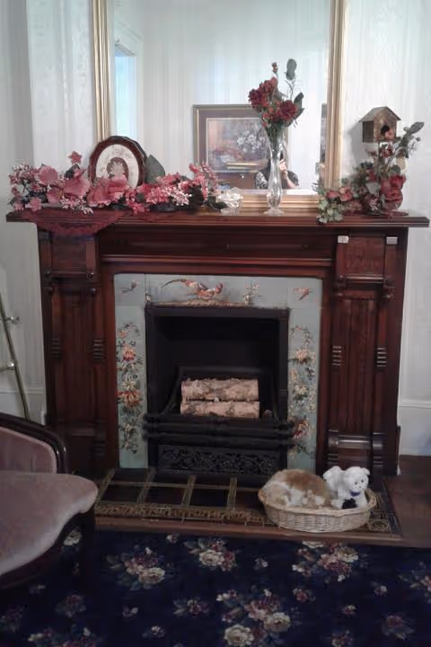 Ornate wooden fireplace mantel with a large mirror, floral arrangements, framed art, and a basket with stuffed animals on a patterned rug.