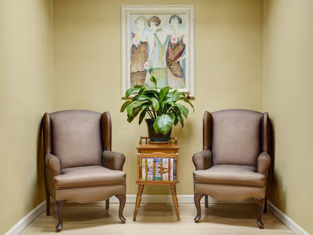 Two brown leather armchairs placed on either side of a small wooden table with a potted green plant on top. Behind the table, a framed painting of three women in vintage-style dresses hangs on a beige wall.