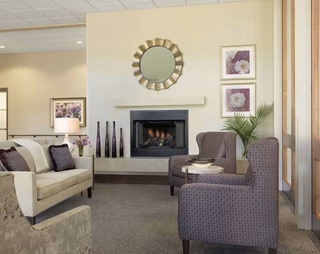 A cozy living room area with a beige sofa adorned with dark purple pillows, two patterned armchairs, a small round side table, and a fireplace with a decorative round mirror above it. The walls are light-colored and decorated with framed floral artwork. There is a potted plant near the armchairs and a lamp on a side table next to the sofa.