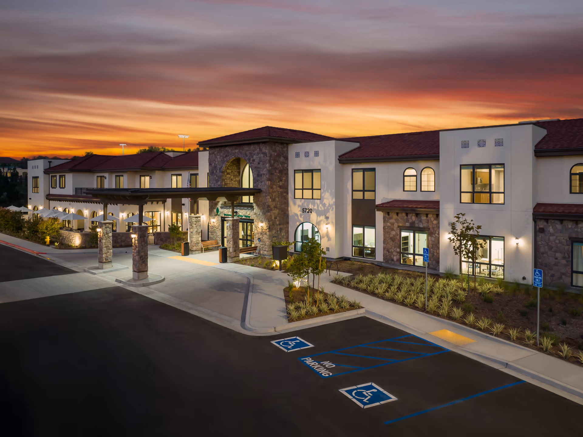 Exterior view of Westmont of Carmel Valley senior living facility at sunset, showing a two-story building with stone and stucco facade, arched windows, a covered entrance, landscaped grounds, and handicap parking spaces in the foreground.