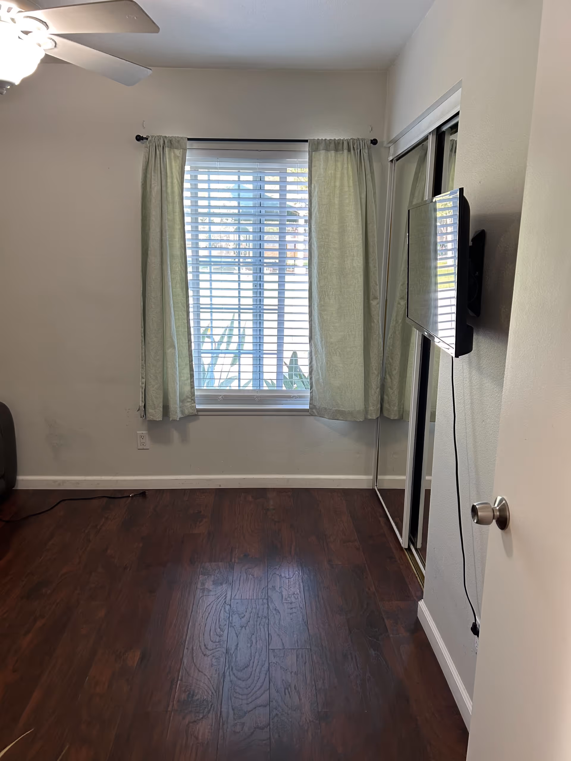 Empty room with dark wooden floor, a window with light green curtains and white blinds, a wall-mounted TV with visible cables, a mirrored closet door, and a ceiling fan with light.