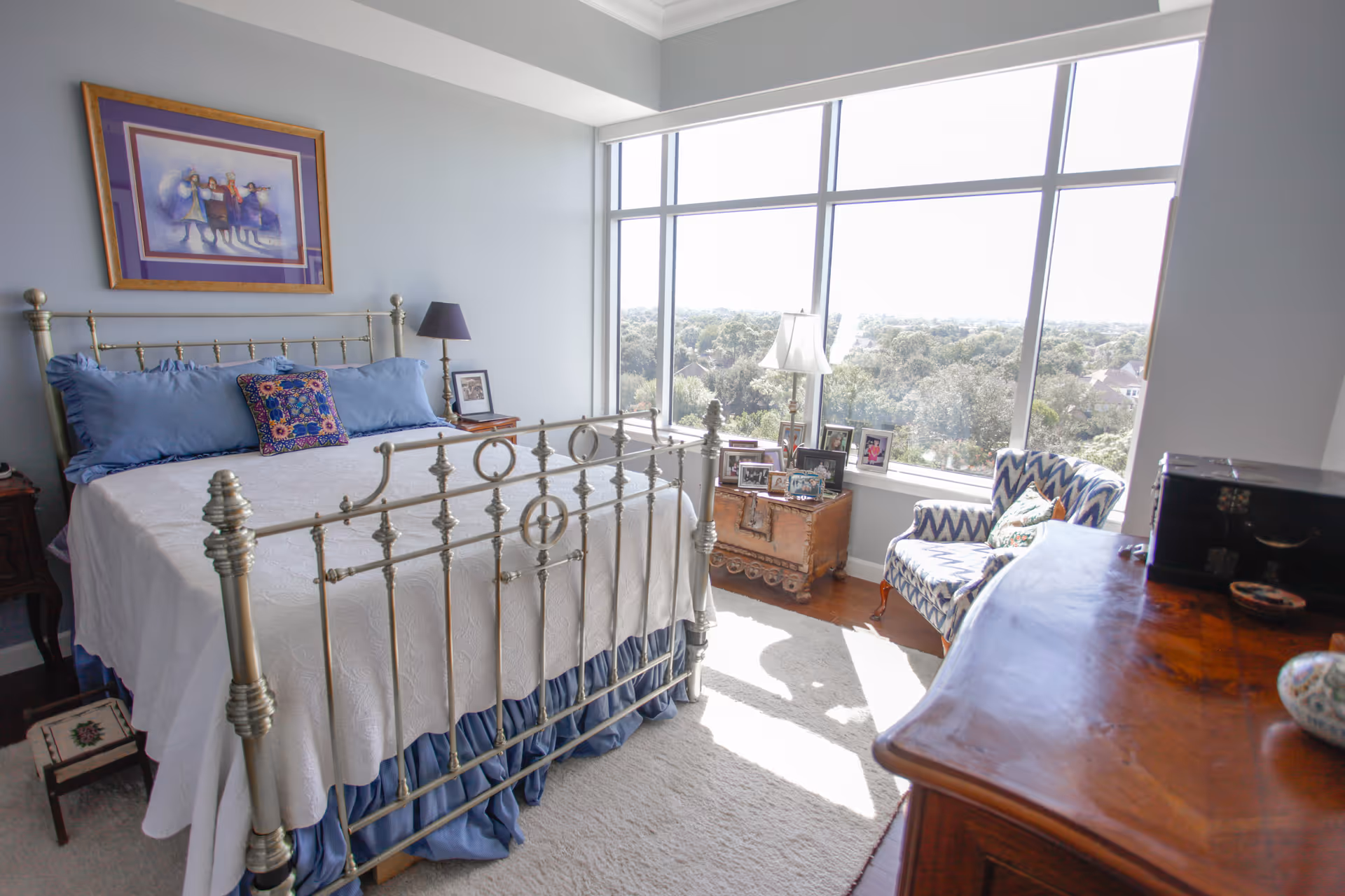 A bright bedroom with a metal bed frame, white bedspread, and blue pillows. There is a framed picture hanging on the wall above the bed. Next to the bed is a small wooden nightstand with a lamp and a framed photo. A large window fills the room with natural light and offers a view of trees outside. In front of the window is a wooden chest with several framed photos on top and a patterned armchair beside it. A wooden dresser is partially visible in the foreground.