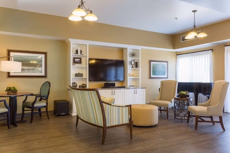 A cozy living room area with a wall-mounted flat-screen TV surrounded by built-in white shelves and cabinets. The room features a striped armchair, two beige upholstered chairs, a round ottoman, a small round table with a plant, and a dining table with chairs. The walls are painted light beige with framed artwork, and large windows with sheer white curtains allow natural light to fill the space.