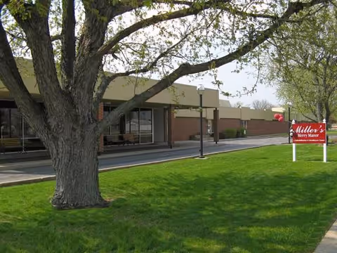 Exterior view of Miller's Merry Manor facility showing a large tree in the foreground, a well-maintained green lawn, a sidewalk, and the building with large windows and a covered entrance. A red sign with white text reads 'Miller's Merry Manor'.