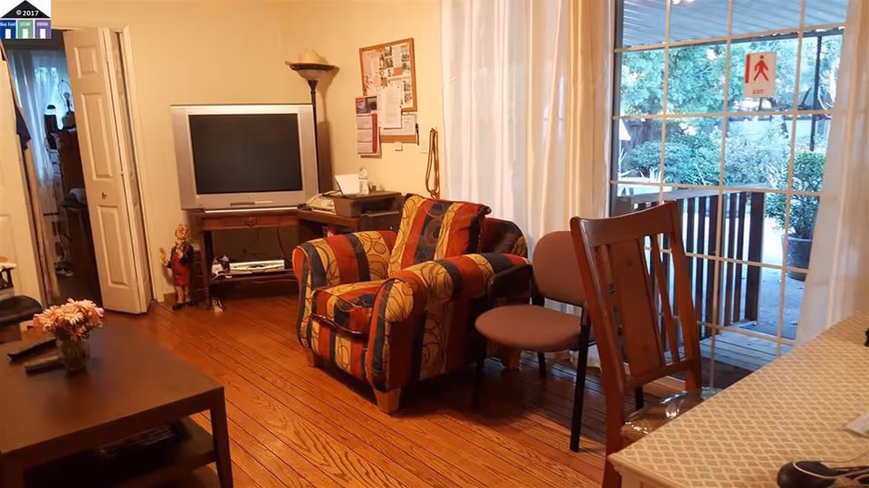 A cozy living room with wooden flooring featuring a colorful patterned armchair, a small round chair, a wooden chair, and a coffee table with a flower vase. There is a TV on a stand against the wall, a floor lamp, and a bulletin board. Large glass sliding doors with white curtains open to an outdoor area with greenery visible.
