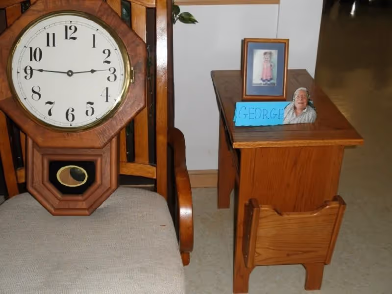 A wooden grandfather clock sits beside a small side table with a framed photo and a blue name card in an indoor senior living setting.