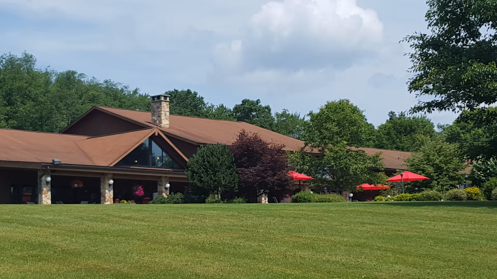 Exterior view of a single-story building with a brown roof and stone pillars, surrounded by green trees and a large grassy lawn. Several red umbrellas are visible on the patio area in front of the building under a partly cloudy sky.