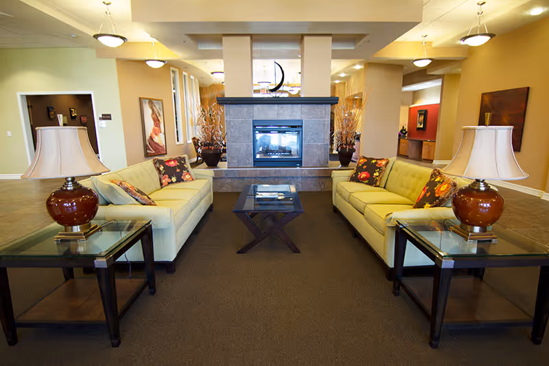 Spacious senior living lounge with two sofas facing a central double-sided fireplace, glass-topped side tables with lamps, and decorative accents.