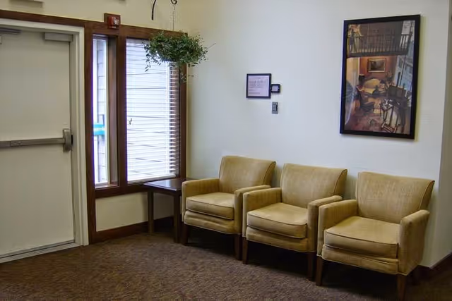 A small waiting area with three beige armchairs lined up against a white wall. There is a framed picture hanging above the chairs and a small table next to a window with blinds. A hanging plant is suspended near the window, and a closed door is visible to the left.
