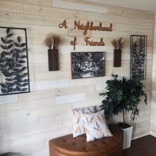 Interior wall decorated with two framed botanical prints, two wall-mounted vases with dried plants, a framed black and white photo, and wooden letters spelling 'A Neighborhood of Friends'. Below the wall decor is a brown cushioned bench with two patterned pillows and a potted green plant in a white planter.