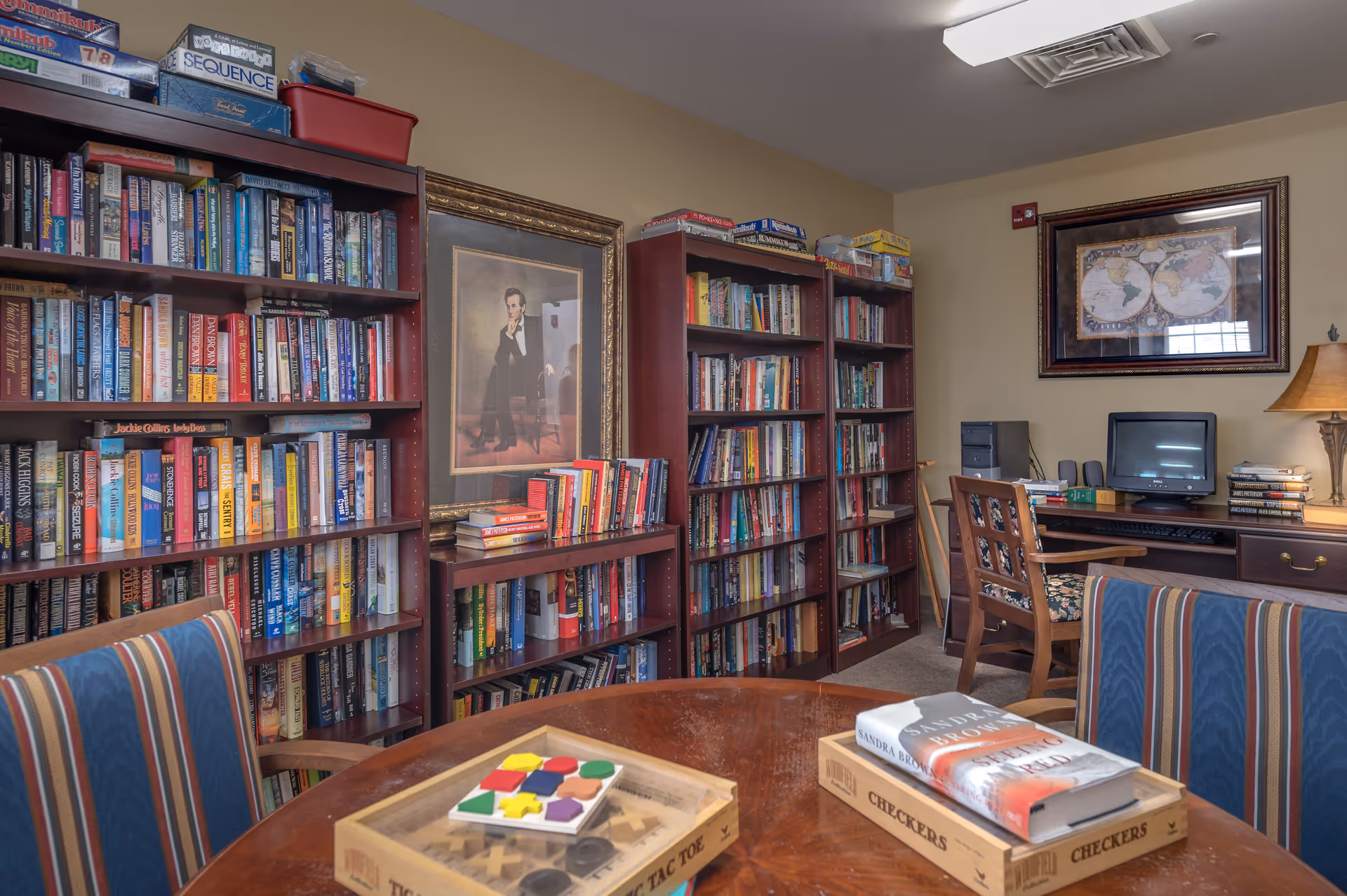 Bookshelves line a cozy common room with a table holding board games and chairs and a computer desk against the wall.