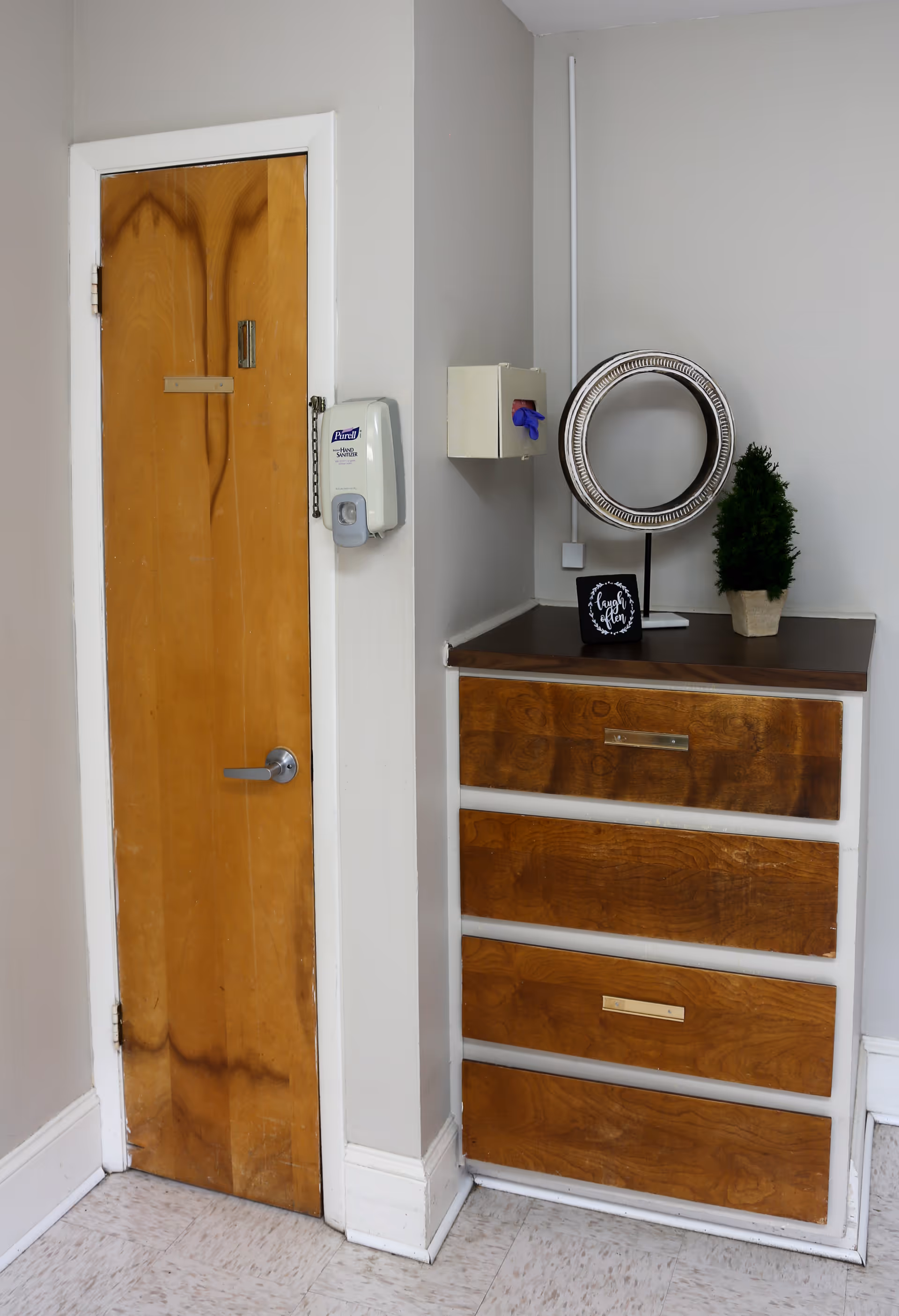 A corner of a room with a wooden door on the left and a wooden chest of drawers on the right. On the wall next to the door is a mounted hand sanitizer dispenser and a small box with a blue glove. On top of the chest of drawers is a decorative round mirror, a small potted plant, and a small sign with the words 'laugh often'. The walls are painted light gray and the floor has light-colored tiles.