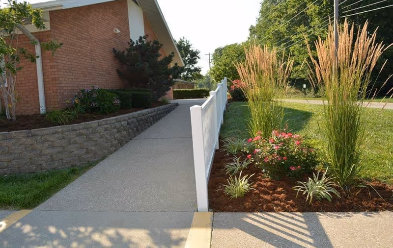 Concrete accessibility ramp with a white railing leading to a brick building, bordered by landscaped flowerbeds and tall ornamental grasses.