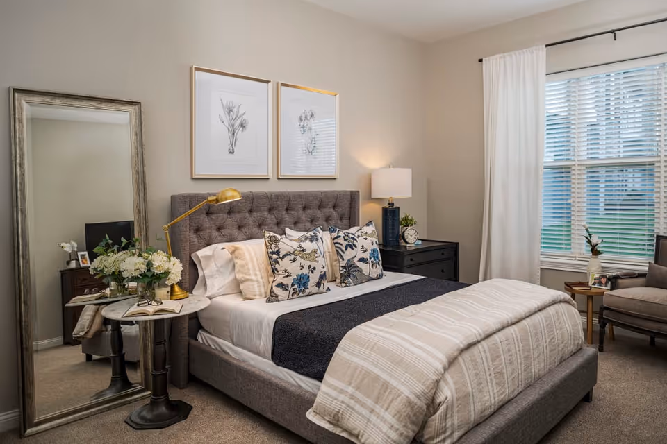 A cozy bedroom with a tufted gray upholstered bed featuring white and floral pillows, a beige striped comforter, and a dark blue throw blanket. There is a round side table with a brass lamp and a vase of white flowers next to the bed. A large floor mirror leans against the wall, reflecting part of the room. Two framed botanical prints hang above the bed. A window with white curtains lets in natural light, and a comfortable armchair with a small side table is placed near the window.