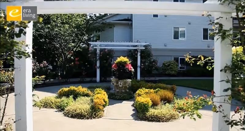 Outdoor garden area with a circular walkway surrounding a central planter filled with colorful flowers and greenery. The garden is framed by white pergola structures and is situated in front of a light-colored building with multiple windows. Trees and shrubs surround the garden area.