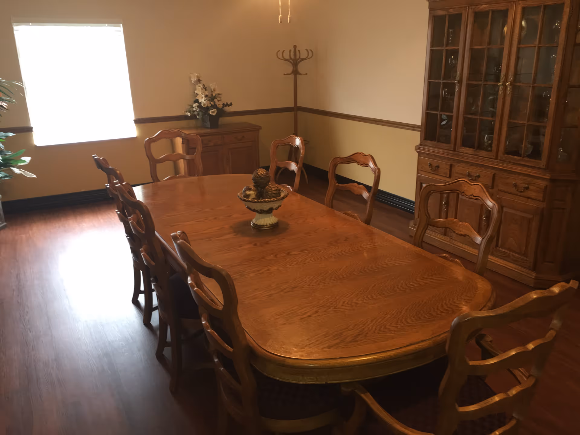 A dining room with a large wooden table surrounded by eight matching wooden chairs. There is a decorative centerpiece on the table. The room has wooden flooring, a window with blinds, a wooden cabinet with glass doors, a sideboard with a floral arrangement, and a wooden coat rack in the corner.