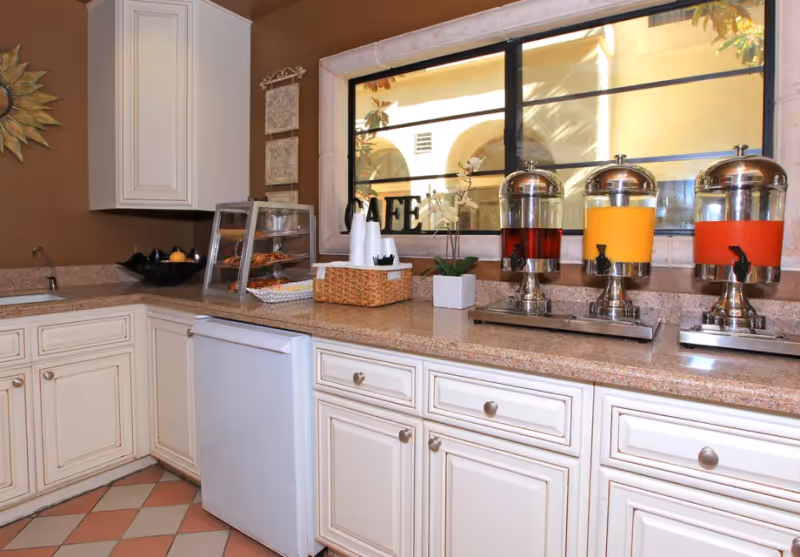 A kitchen counter with three beverage dispensers containing red, orange, and yellow drinks. There is a small refrigerator under the counter, a display case with pastries, a basket with disposable cups, and a small potted plant. The wall is brown with white cabinets and a window showing an outdoor courtyard.