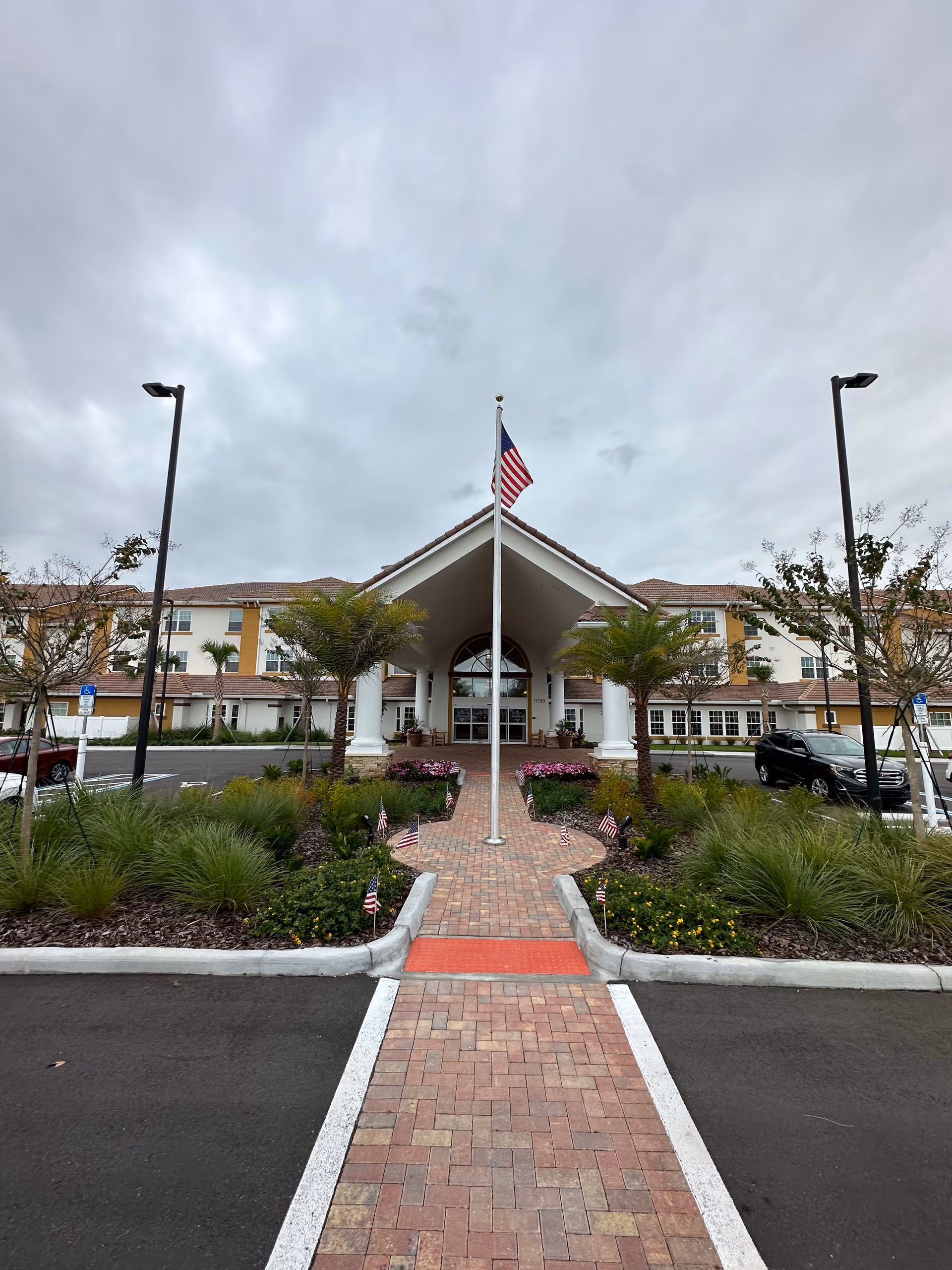 Front exterior view of Pebble Springs Retirement Community building with a brick walkway leading to the entrance, flanked by landscaped greenery and small American flags. A flagpole with an American flag stands in the center of the walkway. The building has a covered entrance with white columns and a tiled roof under a cloudy sky.