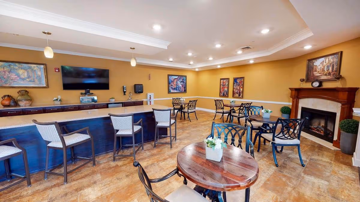 Well-lit common dining area with round tables and chairs, a long counter with barstools, a wall-mounted TV, and a fireplace.