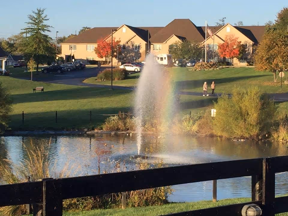 A scenic outdoor view of Liberty Ridge Senior Living featuring a pond with a water fountain creating a mist and a faint rainbow. In the background, there are beige buildings with brown roofs, trees with autumn foliage, a parking lot with cars, and two people walking on a path. The foreground includes a black wooden fence and green grass.
