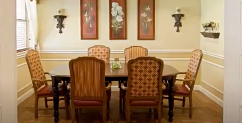 A dining room with a rectangular wooden table surrounded by six upholstered chairs. The walls are painted light yellow with white trim and decorated with three floral paintings and two wall sconces. A window with white blinds and curtains is on the left side, and a small shelf with decorative items is on the right wall.