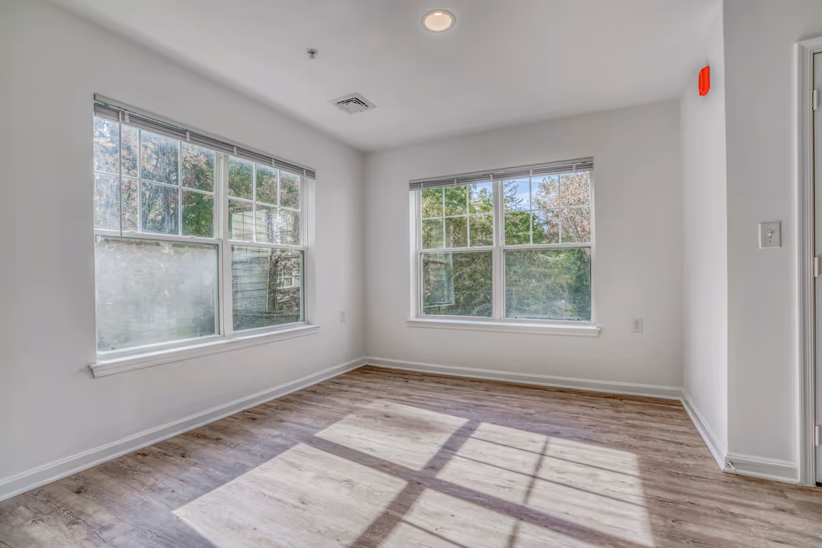 Empty bright room with two large windows, white walls, and wood-look flooring.
