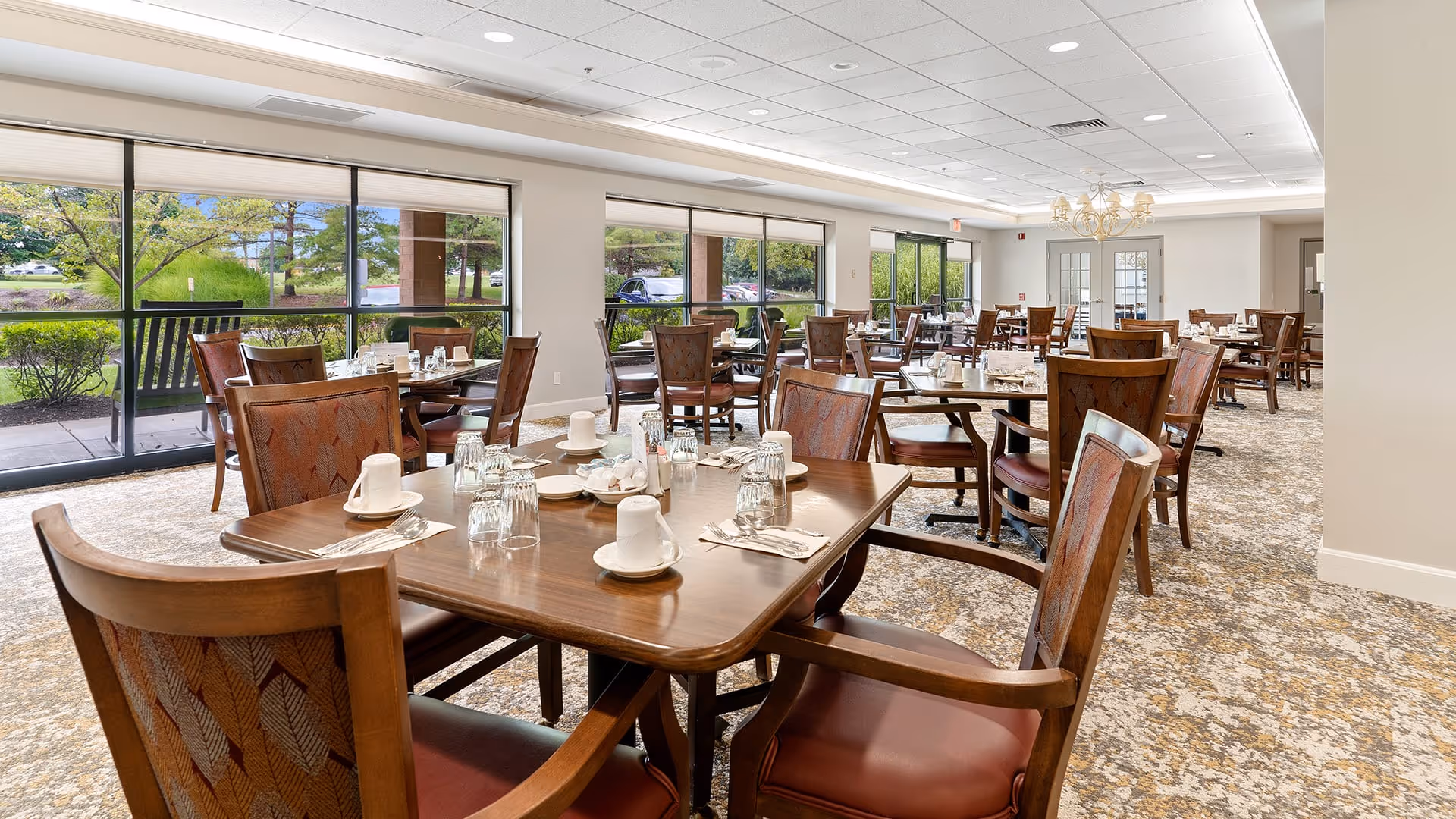 A spacious dining room with multiple wooden tables and chairs arranged neatly. Each table is set with white cups, saucers, glasses, and silverware. Large windows along one side provide a view of greenery and a parking lot outside, allowing natural light to fill the room. The carpet has a patterned design, and the ceiling features recessed lighting and a chandelier.