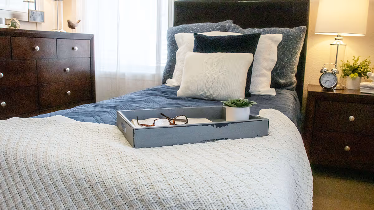 A neatly made bed with layered pillows and a tray holding glasses and a small plant, flanked by dark wooden nightstands and a dresser in a sunlit bedroom.