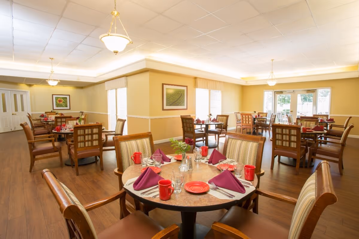 A bright and spacious dining room with multiple round tables set with red mugs, purple napkins, plates, glasses, and silverware. The room has wooden floors, beige walls with white trim, framed artwork, and large windows letting in natural light. Comfortable wooden chairs with cushioned seats surround each table.