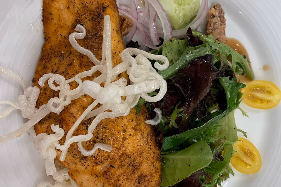 Close-up of a plated cooked fish fillet topped with crispy rice noodles alongside a mixed green salad and halved yellow cherry tomatoes.