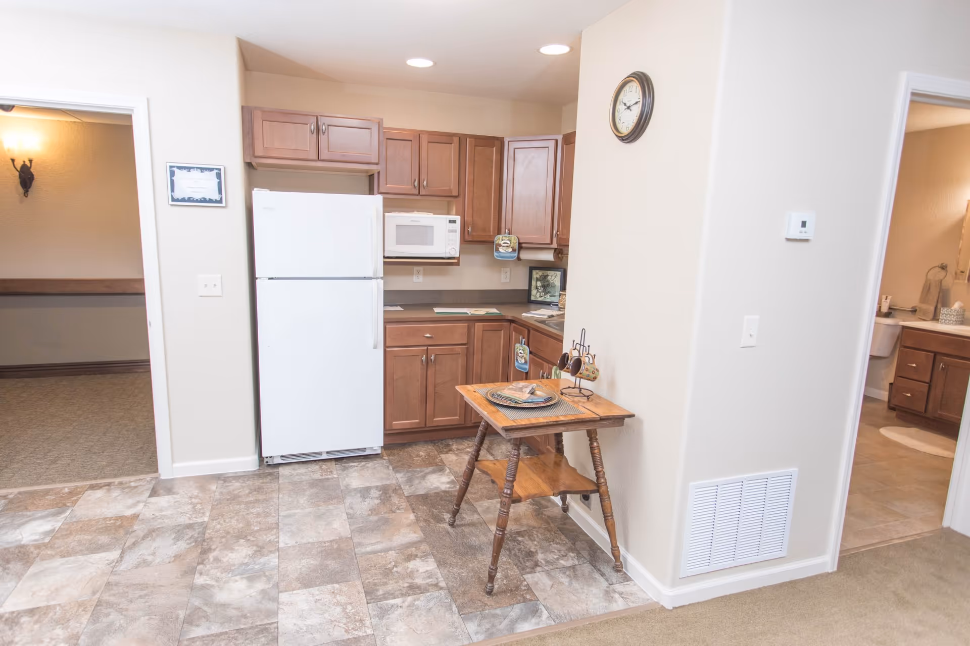 Small kitchen area featuring a white refrigerator and microwave, wooden cabinets, and a small wooden table against a light-colored wall.