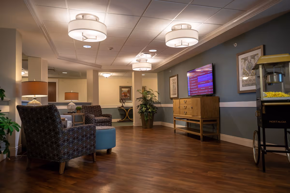 A cozy senior living facility common area with patterned armchairs, a blue ottoman, wooden flooring, and warm lighting from ceiling fixtures and table lamps. A wooden cabinet with a flat-screen TV mounted above it is on the right wall, next to a popcorn machine. Decorative framed artwork and a potted plant add to the inviting atmosphere.