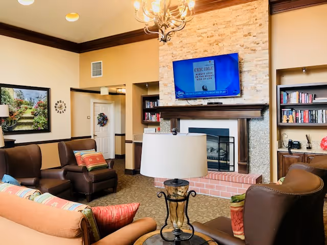A cozy living room area in a senior living facility with comfortable brown leather armchairs and a sofa adorned with colorful pillows. A decorative lamp sits on a small round table in the center. The room features a stone fireplace with a mounted flat-screen TV above it, built-in bookshelves filled with books, and warm beige walls with dark wood trim. A door with a wreath and a framed picture are visible in the background.