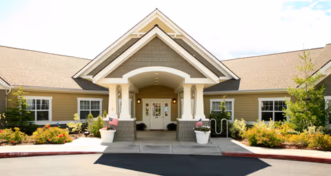 Front entrance of a single-story care facility featuring a covered porch with columns, double doors, landscaping, and American flags.