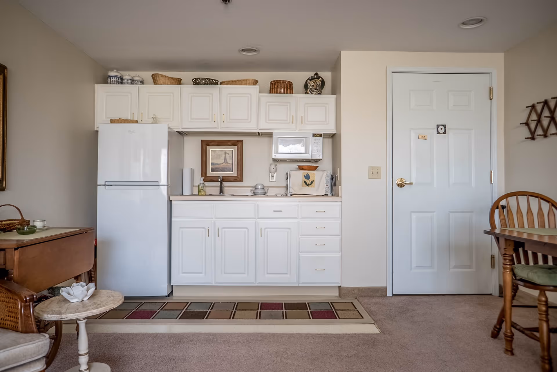 A small kitchen area in a retirement community featuring white cabinets, a white refrigerator, a microwave, a toaster oven, and a sink. There are decorative baskets and jars on top of the cabinets, a framed picture on the wall, and a colorful rectangular rug on the floor. To the right is a white door with a gold handle, and part of a wooden dining table and chair are visible.