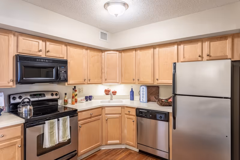 A modern kitchen with light wood cabinets, a stainless steel refrigerator, dishwasher, and stove with a microwave above it. The countertops are white, and there are decorative items including a basket, a blue bottle, and two red apple-shaped containers. A tea kettle sits on the stove, and two striped kitchen towels hang from the oven handle.
