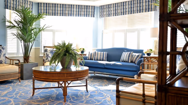 Sunlit sitting room with a blue sofa, wicker armchairs, a round wooden coffee table topped with a fern, and a patterned carpet.