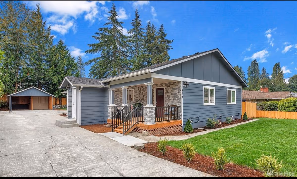 Exterior view of a single-story gray house with a small covered front porch, stone accents, and a well-maintained lawn. There is a driveway leading to a detached garage in the background, surrounded by tall trees and a wooden fence.
