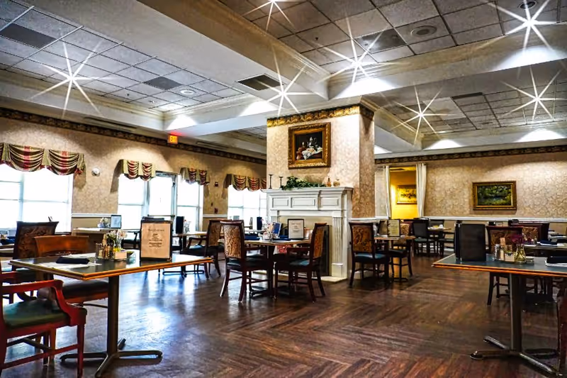 A spacious dining room with multiple tables and chairs arranged neatly. The room features a decorative fireplace with a framed painting above it, patterned wallpaper, and large windows with valance curtains allowing natural light to enter. The ceiling has recessed lighting and a grid pattern, and the floor is wooden.