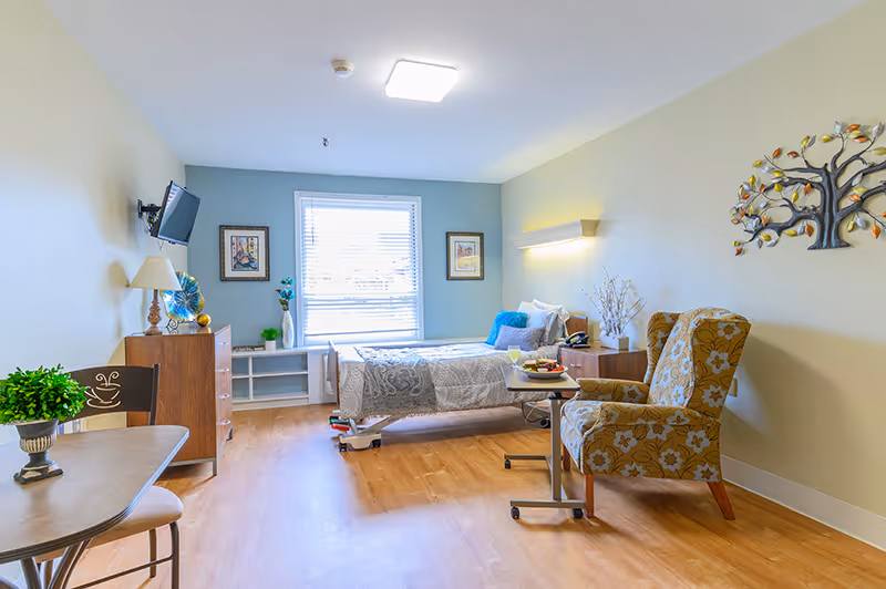 A bright and clean senior living bedroom with a single bed covered in a gray and white patterned blanket. Next to the bed is a wooden nightstand with a lamp and a telephone. A comfortable floral armchair and a small rolling table are positioned near the bed. The room has light-colored walls, a window with blinds letting in natural light, a wall-mounted TV, a wooden dresser, and decorative wall art including a metal tree sculpture and framed pictures. There is also a small table with a plant and chair in the foreground.