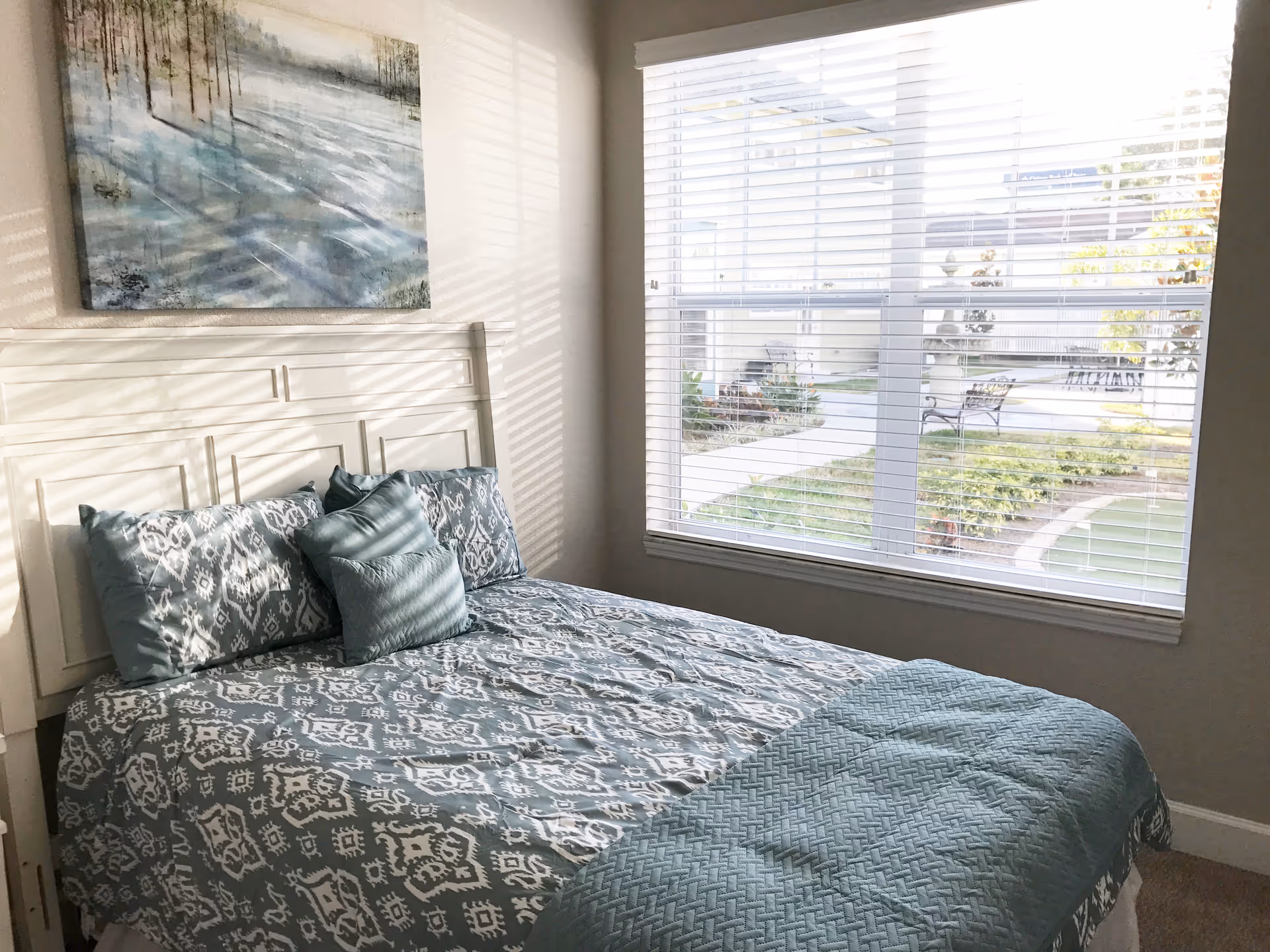 Sunlit bedroom with a teal-patterned bed, white headboard, artwork above, and a large window overlooking a courtyard.