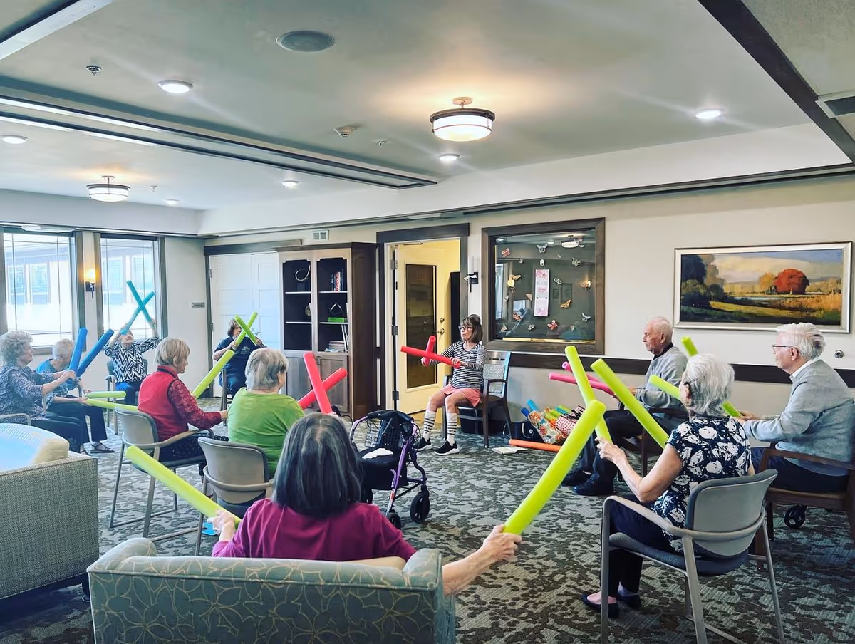 A group of elderly people seated in a circle in a well-lit common room, participating in a group exercise using colorful foam sticks led by an instructor. The room has comfortable chairs, carpeted floor, and wall decorations including a painting and a bulletin board.