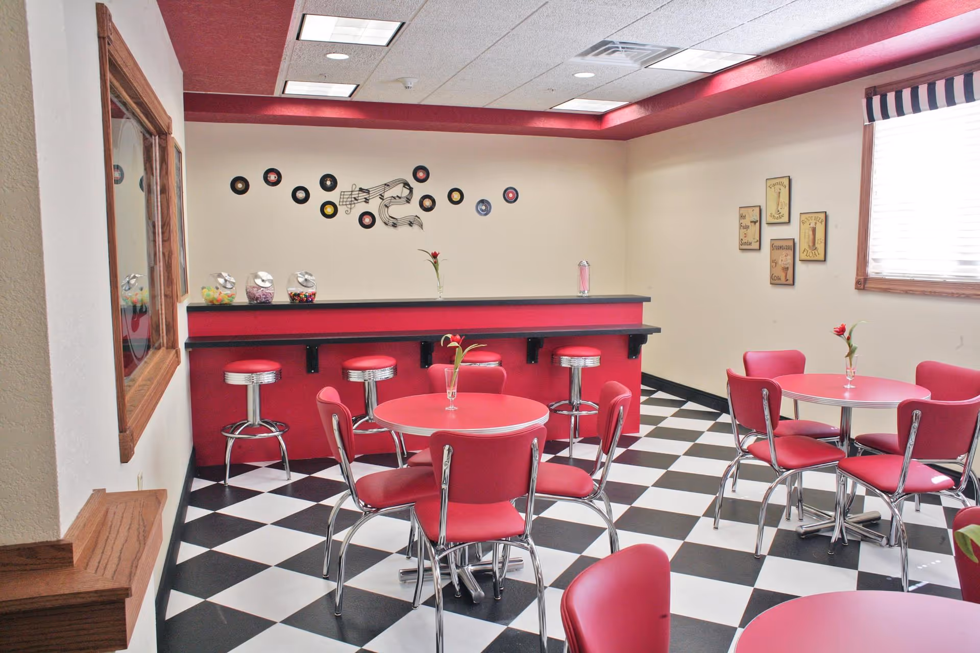 A retro-style dining area with black and white checkered floor tiles, red tables and chairs, and a red counter with chrome bar stools. The walls are decorated with vinyl records and musical notes, and there are small vases with red flowers on the tables. A window with striped valance allows natural light into the room.